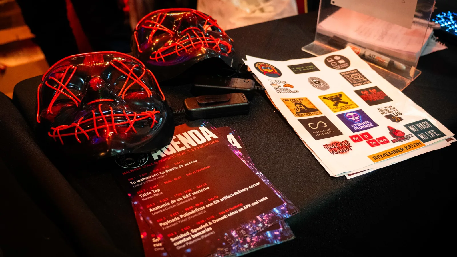 Máscaras con luces LED rojas, stickers y la agenda de charlas sobre una mesa del stand
